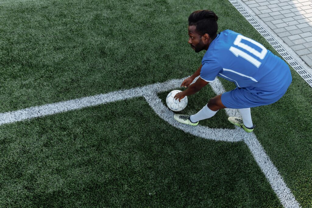 A soccer player in a blue jersey prepares to kick the ball from the corner on an artificial turf field.